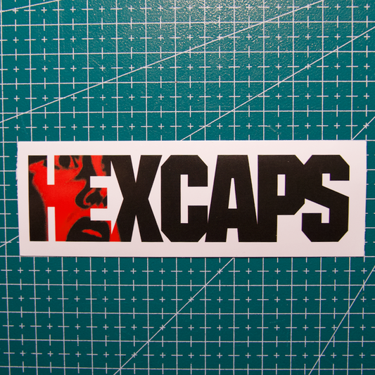 A rectangular white sticker or decal with the word 'HEXCAPS' in bold black letters, where the letter H is filled with a red graphic, placed on a green cutting mat background.
